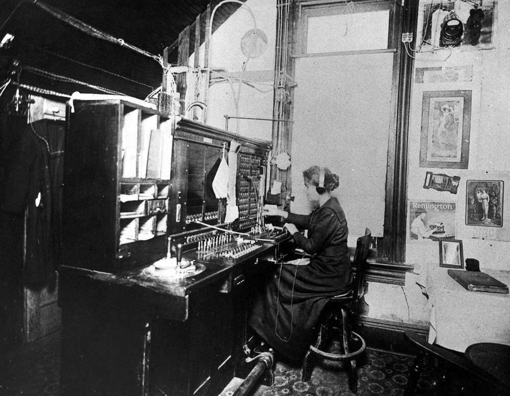 A telephone exchange operator at a manual switchboard, c. 1900 — paired connections through a network of patch cords.