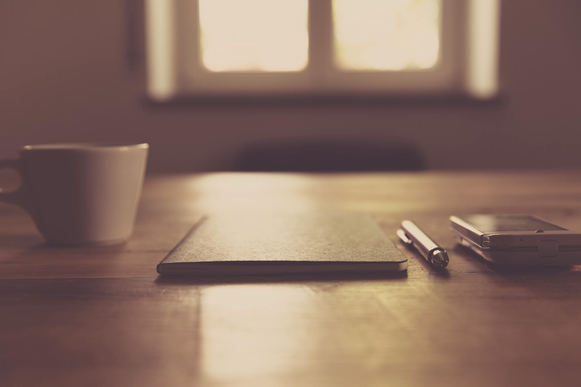 A quiet desk in soft window light: a notebook, a pen, a phone and a cup of coffee